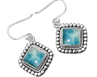 Pode incluir: Brincos de prata com um pendente quadrado de pedra azul e branca, engastado em uma moldura de prata com uma borda de contas.