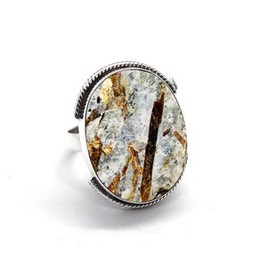 Op de afbeelding: Een zilveren ring met een grote ovale edelsteen. De edelsteen is een mix van witte, bruine en gouden kleuren. De ring heeft een getextureerde zilveren band.