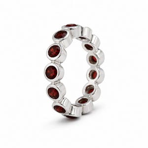 May include: A silver eternity band ring featuring a continuous row of round, deep red garnet gemstones set in individual polished metal bezels. The ring displays a uniform circular design with each stone securely encased in a smooth silver setting.