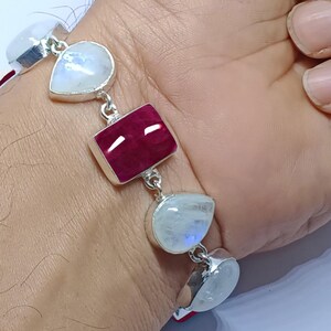 May include: A silver bracelet featuring a rectangular ruby gemstone and teardrop-shaped moonstones. The bracelet is connected by silver links and is worn on a wrist. The gemstones have a polished finish.