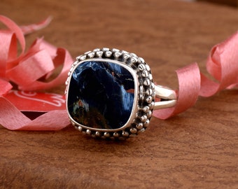 May include: A silver ring featuring a square, dark blue gemstone with a marbled pattern of lighter blue and brown. The ring has a detailed setting of small, round silver beads. The ring is a piece of jewellery.