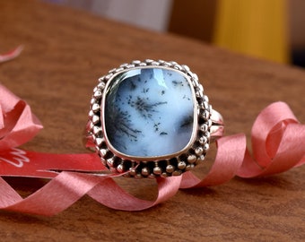 Può includere: Un anello d'argento con una pietra preziosa di agata dendritica di forma quadrata. L'anello ha un bordo perlato ed è posto su una superficie di legno con un nastro rosa.
