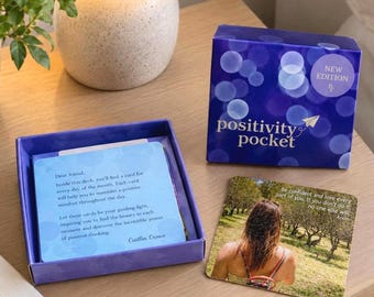 Puede incluir: Un juego de cartas Positivity Pocket en una caja azul con patrones de luces bokeh. La tapa presenta el texto New Edition y Positivity Pocket. Dentro, una tarjeta azul claro muestra un texto de Caitlin Crowe, y una tarjeta cuadrada muestra a una persona en un campo con la cita Be confident and love every part of you.