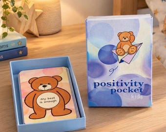 Puede incluir: Un mazo de cartas en una caja azul claro titulada Positivity Pocket Kids, con una ilustración de un oso de peluche marrón. La tapa muestra un oso en un avión de papel, y la caja abierta muestra una carta con un oso y el texto My best is enough.