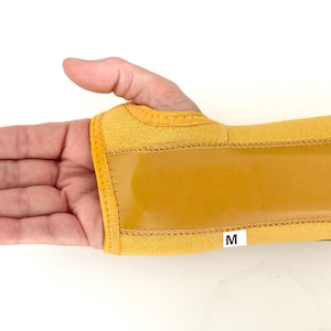 May include: A yellow wrist support with a tan-coloured, rectangular support. The brace is secured with a hook and loop closure and features a small white tag with the letter "M".