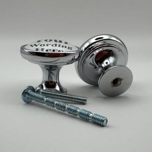 May include: Two chrome cabinet knobs with the text "Your Wording Here" on the top. The knobs are round with a decorative base and come with silver screws for installation. The knobs are on a white background.