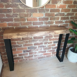 May include: A rustic wooden console table with a live edge top and black metal hairpin legs. The table is set against a brick wall and a potted plant is on the floor.