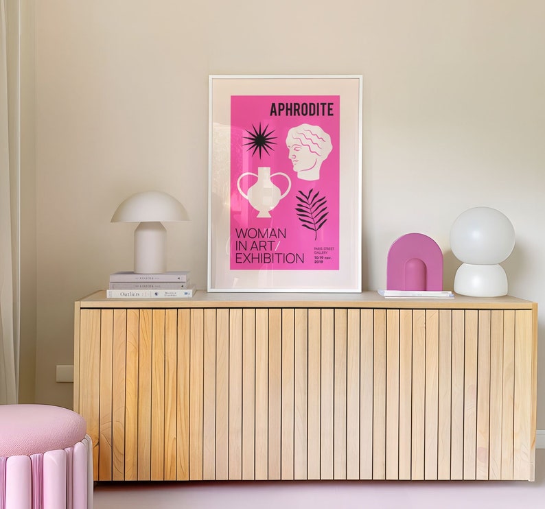 以下が含まれることがあります： ピンクのポスターに「APHRODITE WOMAN IN ART EXHIBITION」と書かれた明るい木製のキャビネット。白い花瓶には黒い星と女性の顔があります。キャビネットの上には、白いシェードのランプと透明なガラスシェードのランプの2つのランプがあります。キャビネットの上には、ピンクのアーチ型のオブジェクトがあります。