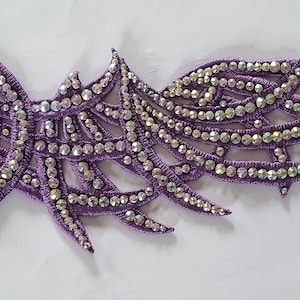May include: A decorative purple applique with an abstract, swirling design. The applique is adorned with many iridescent rhinestones, creating a sparkling effect. The design features pointed, wing-like shapes.
