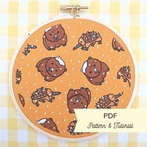 May include: A round embroidery hoop with a yellow fabric background featuring a repeating pattern of embroidered echidnas and wombats. The text "PDF Pattern & Tutorial" is printed on a white banner at the bottom of the hoop.
