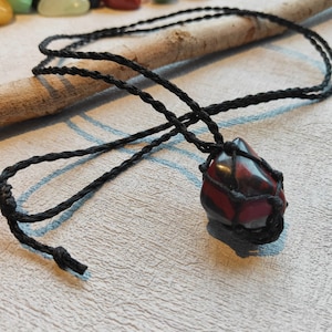 May include: A black cord necklace with a red and black gemstone pendant wrapped in a macrame knot.