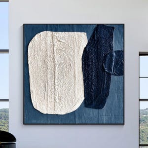 Puede incluir: Una pintura abstracta cuadrada que presenta una gran forma orgánica blanca texturizada junto a una forma vertical azul marino oscuro sobre un fondo azul profundo. La obra se exhibe en una pared blanca sobre un sofá capitoné azul marino.