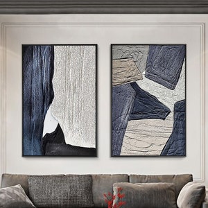 May include: Two abstract paintings in black frames, one with white and blue geometric shapes, the other with white and gray geometric shapes. The paintings are hanging on a white wall above a gray sectional sofa.