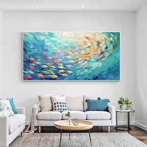 May include: A large abstract painting of a school of colorful fish swimming in a turquoise blue ocean. The painting is framed and hanging on a white wall in a living room setting.