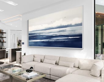 May include: Large abstract painting depicting a seascape with shades of blue and white. The artwork features broad brushstrokes suggesting waves and sky, with a few birds in flight. The painting is mounted on a white wall above a beige sectional sofa.