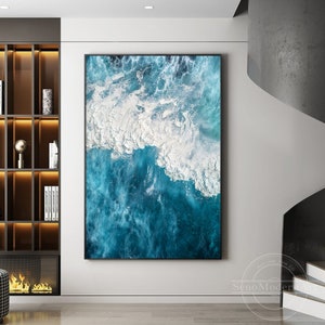 May include: An abstract painting of a blue ocean with white foamy waves crashing on the surface. The painting is framed in a black frame and is hanging on a white wall.