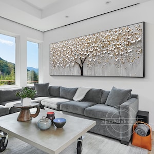 May include: A large abstract painting of a white tree with gold accents hanging on a wall in a living room. The painting is above a gray sectional sofa with a white throw blanket. A coffee table with a gray top and black wheels sits in front of the sofa.