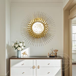 Puede incluir: Un espejo dorado con forma de sol con un espejo redondo central y detalles blancos en forma de pétalos. El espejo está montado en una pared blanca sobre un mueble blanco con herrajes dorados. Un jarrón con flores blancas y una figura dorada están sobre el mueble.
