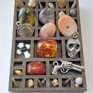 May include: A rectangular wooden grid organiser containing various small items including polished stones, seashells, a silver-toned skull charm, a silver-toned revolver charm, a white cross pendant, and a round metal charm with Chinese characters.