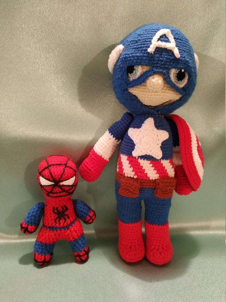 May include: Two crocheted superhero toys. Captain America is wearing a blue and red costume with a white star and a red, white, and blue shield. Spiderman is wearing a red and blue costume with a black spider on the chest.