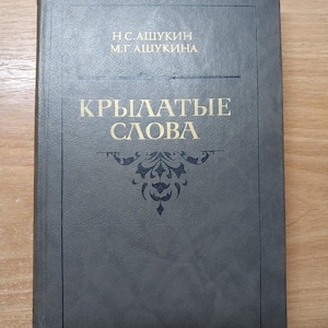 May include: A hardcover book with a dark gray cover. The title "КРЫЛАТЫЕ СЛОВА" is printed in gold lettering, along with the authors' names. A decorative emblem is centered below the title. The book is on a wooden surface.