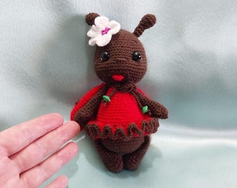 May include: A brown crocheted ladybug doll wearing a red dress with a white flower on its head.