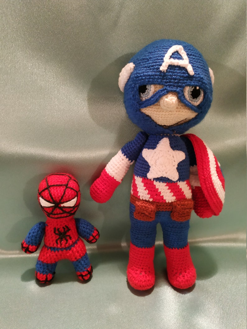 May include: Two crocheted superhero toys. Captain America is wearing a blue, red, and white costume with a red, white, and blue shield. Spiderman is wearing a red and blue costume with a black spider on the chest.