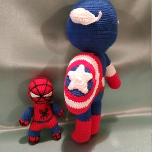 May include: A crocheted Captain America doll with a red, white, and blue costume and a shield. A smaller crocheted Spiderman doll is standing next to Captain America.