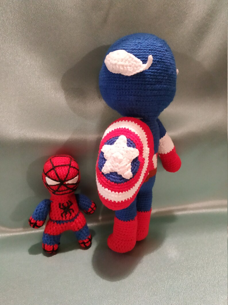 May include: A crocheted Captain America doll with a red, white, and blue costume and a shield. A smaller crocheted Spiderman doll is standing next to Captain America.