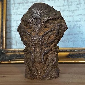 Könnte beinhalten: Eine detaillierte Bronzeskulptur einer Kreatur mit einer strukturierten Oberfläche und einem abgerundeten Kopf. Die Skulptur ist dunkelbraun und hat ein detailliertes Gesicht.