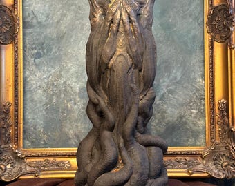 May include: A dark gray sculpture of a humanoid figure with a textured surface. The figure has a detailed face and is surrounded by tentacle-like appendages. The sculpture is set against a backdrop of a gold-framed mirror and a brown cloth.