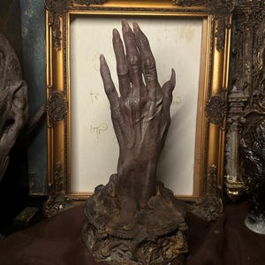 Puede incluir: Una escultura oscura y desgastada de una mano con uñas largas y puntiagudas. La mano es de un color marrón oscuro y parece emerger de una base texturizada y ornamentada. El fondo incluye un cuadro con marco dorado y otros elementos decorativos.
