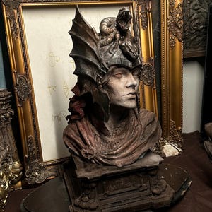 May include: A detailed bust sculpture of a figure wearing a helmet adorned with a coiled snake. The figure has a weathered, textured appearance in shades of brown and grey, draped in a cloak, and mounted on a rectangular pedestal base.