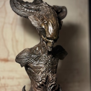 Puede incluir: Un busto esculpido detallado de una criatura con una textura de piel gris-marrón. La criatura tiene una cabeza grande y puntiaguda con un cuello largo y delgado y una pequeña boca abierta. Los ojos de la criatura están cerrados y su cuerpo está cubierto de intrincados detalles.