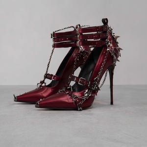 Puede incluir: Un par de zapatos de tacón alto en color rojo metálico profundo con punta afilada, múltiples correas con hebillas en el tobillo y adornos de cadenas plateadas. Los tacones presentan púas plateadas afiladas en la parte posterior y lateral.
