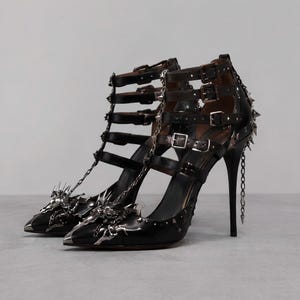 May include: Black high-heeled pumps featuring a pointed toe, multiple buckled ankle straps, and silver-toned metallic embellishments. The design includes intricate spiked hardware on the toe and heel, along with delicate hanging chains.
