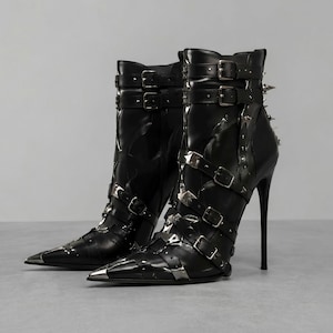May include: A pair of black ankle boots featuring a pointed toe with a silver metal cap, a high stiletto heel, and multiple adjustable straps with silver buckles and studs wrapping around the shaft and foot, including spikes on the back heel.