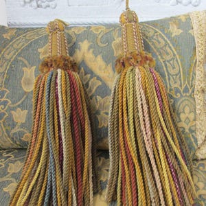 Puede incluir: Dos borlas decorativas con una variedad multicolor de cordones trenzados en tonos oro, verde oliva, rojo óxido y azul apagado. Cada borla tiene una sección superior estructurada y envuelta con un pequeño lazo para colgar en la parte superior.