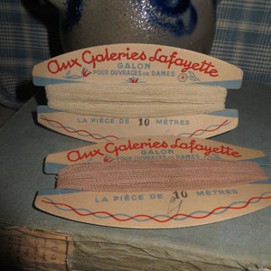 Könnte beinhalten: Zwei Spulen mit weißem und beigefarbenem Besatz mit der Aufschrift "Aux Galeries Lafayette Galon Pour Ouvrages de Dames La Pièce de 10 Mètres".