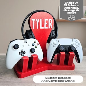May include: A red wooden gaming stand featuring a central vertical support with the name TYLER in white letters, designed to hold one pair of black headphones and two game controllers, with text stating Choice Of 13 Colours. Any Name, Message Or Image.