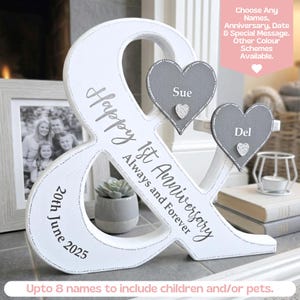 May include: A white wooden ampersand sign featuring the text Happy 1st Anniversary Always and Forever and 20th June 2025. Two grey heart-shaped plaques are attached, labelled Sue and Del, each with a small silver glitter heart accent.
