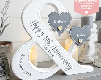 Puede incluir: Un signo ampersand de madera blanca con el texto Happy 11th Anniversary My Love y Established 2015. Dos corazones de madera gris están unidos, cada uno mostrando un nombre, Robert y Jacky, con un pequeño corazón dorado de adorno.