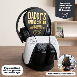 May include: A black gaming stand featuring a central plaque with the gold text DADDY'S GAMING STATION, LOVE FROM KATIE, CHRISTOPHER & CALLUM, and a controller icon. The stand holds black headphones and a white and black game controller.
