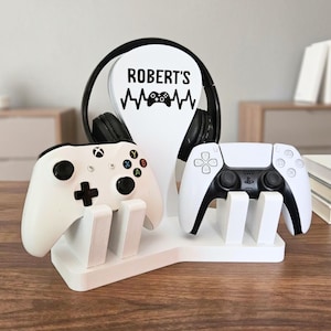 May include: A white gaming accessory stand featuring a central vertical support with the text "ROBERT'S" above a heartbeat line and a game controller icon. The stand holds a black headset and two white game controllers on its base.