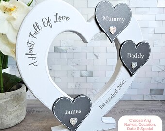 May include: White wooden heart-shaped sign with the text "A Heart Full Of Love" and "Established 2022". Three smaller gray hearts with the names "Mummy", "Daddy", and "James" are attached to the larger heart.