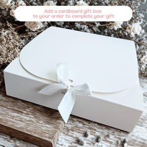 May include: A white cardboard gift box with a rounded flap closure secured by a white ribbon tied in a bow. The box features the text: Add a cardboard gift box to your order to complete your gift. The design is clean, simple, and functional.
