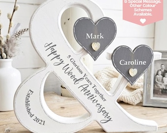 Puede incluir: Un signo ampersand de madera blanca con el texto "5 Glorious Years Together Happy Wood Anniversary" y "Established 2021". Se incluyen dos placas de madera en forma de corazón gris, etiquetadas como "Mark" y "Caroline" con pequeños corazones dorados.