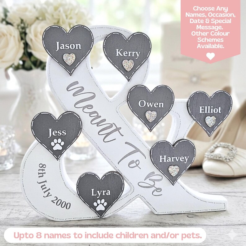 May include: A white ampersand-shaped wooden display featuring the text Meant To Be and 8th July 2000. It includes six dark grey heart-shaped plaques with the names Jason, Kerry, Owen, Elliot, Jess, and Lyra, each accented with a small glittery heart or paw print.