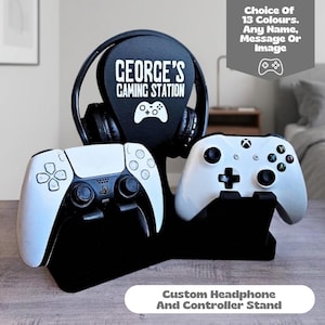 May include: A black gaming stand featuring a central vertical panel with the white text "GEORGE'S GAMING STATION" and a controller icon. The stand holds a pair of black over-ear headphones and two white video game controllers on its base.