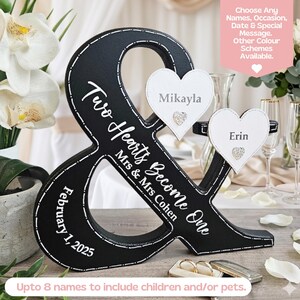 May include: A black ampersand-shaped wooden sign featuring white text that reads Two Hearts Become One, Mrs & Mrs Cohen, and February 1, 2025. Two white heart-shaped attachments are displayed, each featuring a name and a small crystal accent.